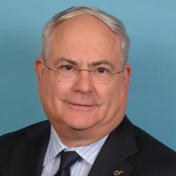 Headshot of a man with light-colored hair and glasses, wearing a dark suit, light-blue striped shirt, and tie.