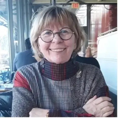 Smiling person with short light hair and round glasses, wearing a patterned collared top, sitting in a restaurant.