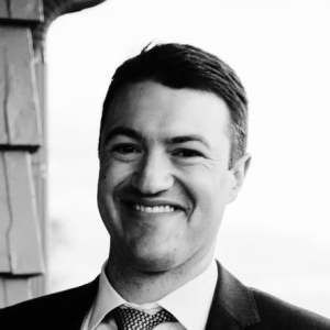 A black-and-white headshot of a smiling person wearing a suit, white shirt, and patterned tie.