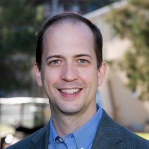 A smiling man in a blue button-down shirt and a grey blazer, shown in a head-and-shoulders portrait outdoors.
