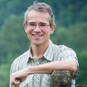 A person with glasses in a plaid shirt smiles, resting an arm on a wooden post against a blurred green background.