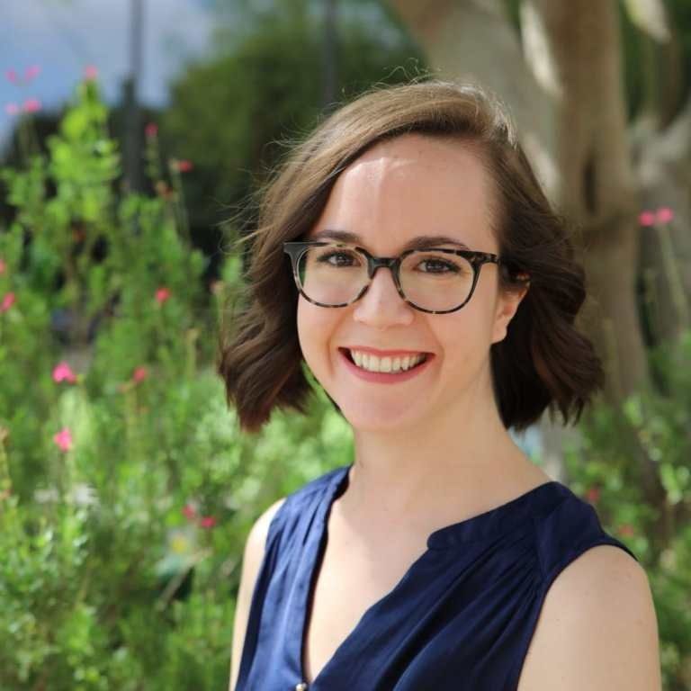 A person with shoulder-length brown hair and glasses smiles in front of a blurry outdoor background of green plants.