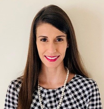 A headshot of a smiling person with long dark hair, wearing a pearl necklace and a patterned top.