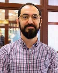 A person with a beard and glasses wearing a purple and white checkered shirt, standing in front of a glass wall.