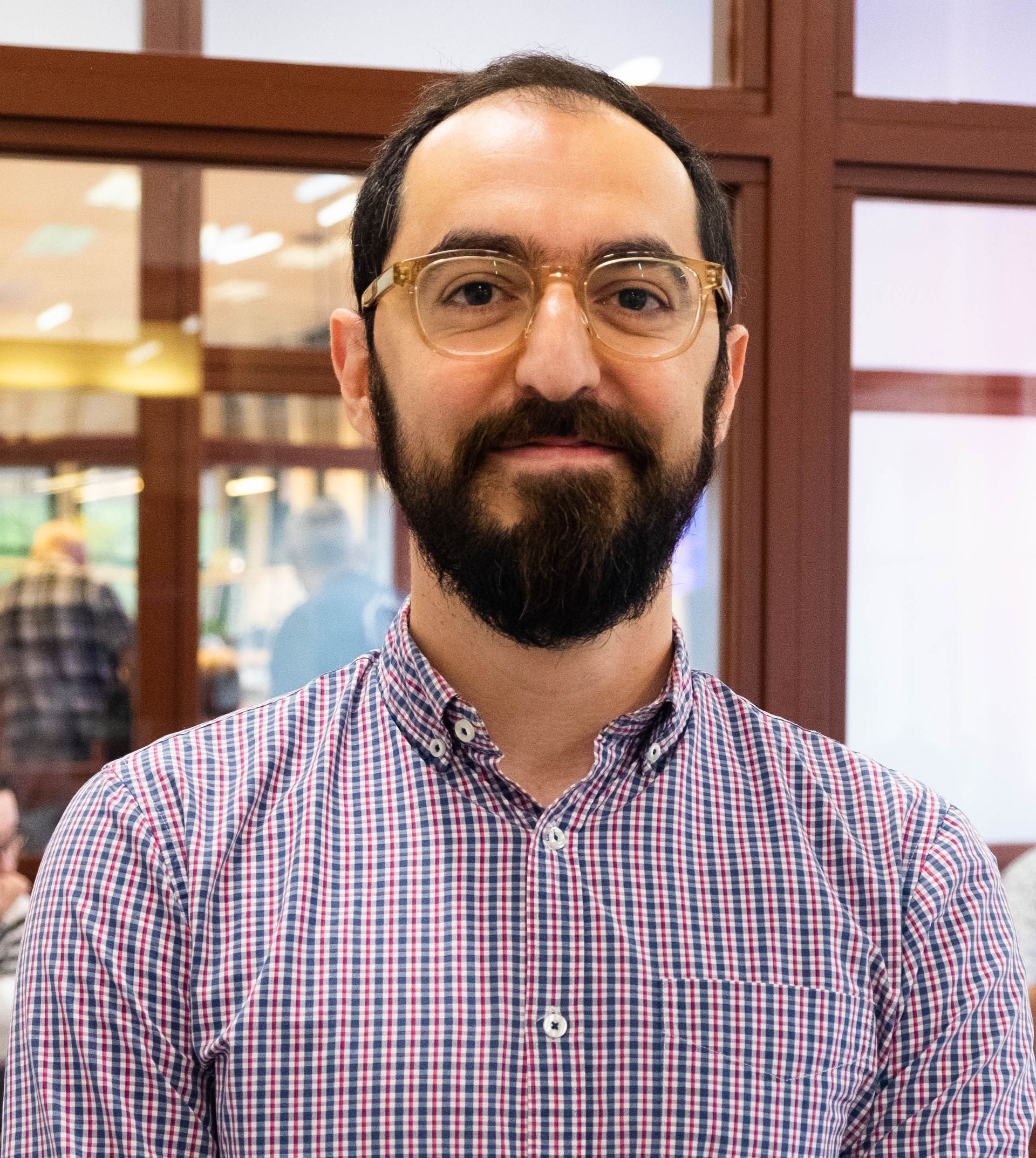 A person with a beard and glasses wearing a purple and white checkered shirt, standing in front of a glass wall.