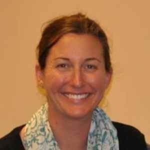 A smiling person with shoulder-length brown hair, wearing a white and teal patterned scarf against a plain wall.