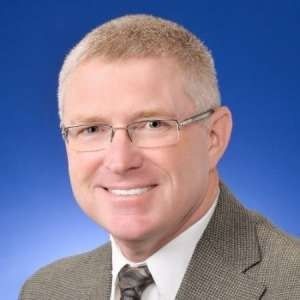 Headshot of a smiling person wearing glasses, a white collared shirt, and a gray blazer against a blue background.