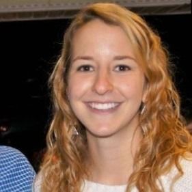 A smiling person with wavy, shoulder-length light brown hair wearing a white top against a dark background.