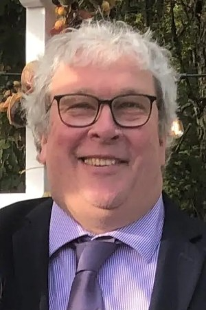 Smiling person with grey hair and glasses, wearing a dark suit jacket, light purple shirt, and tie against an outdoor background.