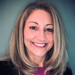A person with shoulder-length, blonde-highlighted hair smiling warmly at the camera against a muted teal background.