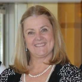 A smiling person with shoulder-length blonde hair, wearing a black and white patterned top and a pearl necklace.