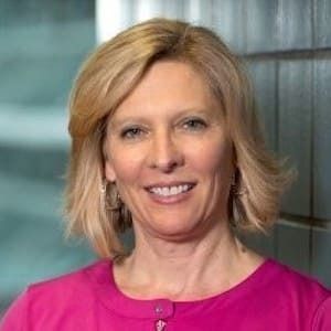 A professional headshot of a person with blonde hair wearing a bright pink collared shirt, smiling against a blurred wall.