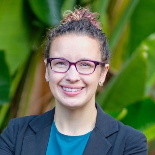 A person with curly hair pulled into a bun and purple glasses smiles against a backdrop of large green leaves.