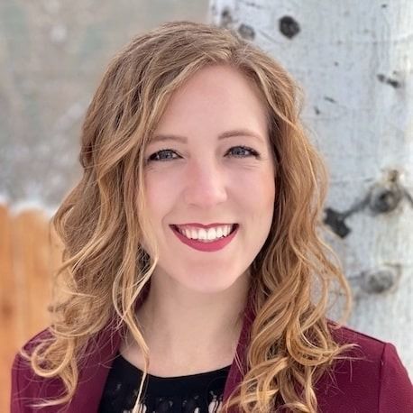 A woman with wavy blonde hair wearing a maroon blazer smiles in front of a tree trunk.