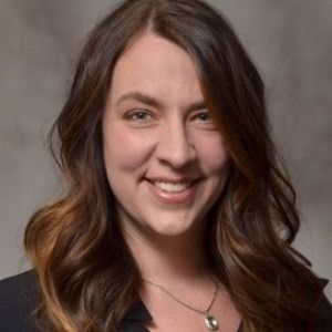 A headshot of a smiling person with long, wavy brown hair, wearing a dark top and a simple silver necklace.