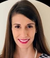 A headshot of a smiling person with long dark hair, wearing a pearl necklace and a patterned top.