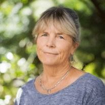 A person with shoulder-length blonde hair and a soft smile wearing a blue top and a silver necklace outdoors.