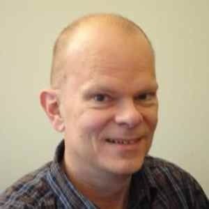 A person with a shaved head and light skin wearing a plaid shirt, smiling slightly against a plain, light background.