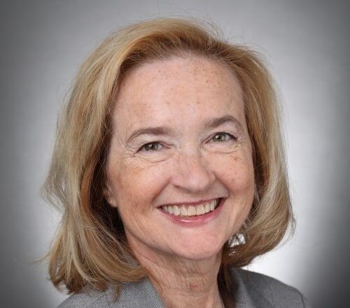 Headshot of a smiling person with shoulder-length blonde hair, wearing a grey blazer against a neutral grey background.