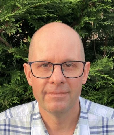A person with a shaved head and glasses wearing a plaid shirt, posed against a backdrop of green evergreen foliage.
