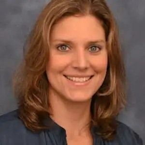 A person with shoulder-length wavy hair smiles toward the camera against a neutral gray background.