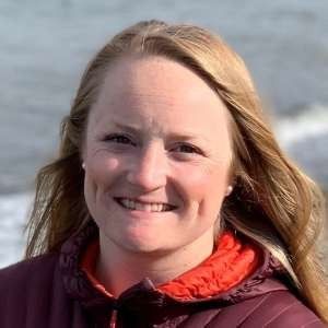 A smiling person with long blonde hair, wearing a maroon jacket with an orange collar, outdoors against a blurred water backdrop.