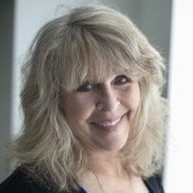 Headshot of a person with shoulder-length, layered blonde hair, wearing a dark top, smiling softly against a light wall.