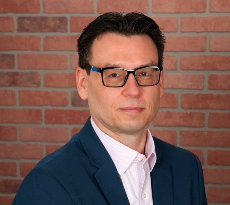 A professional portrait of a person wearing glasses and a blue blazer, standing in front of a brick wall background.