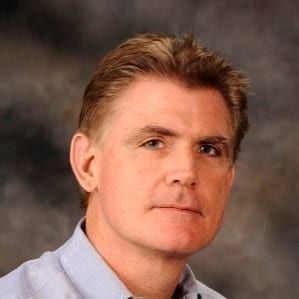 A person with short brown hair wearing a light blue collared shirt, shown in a headshot against a dark, mottled background.