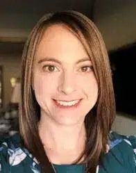 A smiling person with shoulder-length brown hair, wearing a dark green floral-patterned top.