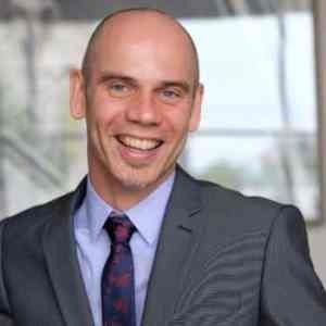 A person with a bald head wearing a grey suit, light blue collared shirt, and patterned tie, smiling against a blurred background.
