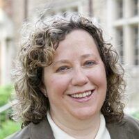 A person with curly hair smiling warmly at the camera, wearing a dark jacket over a white collared shirt.