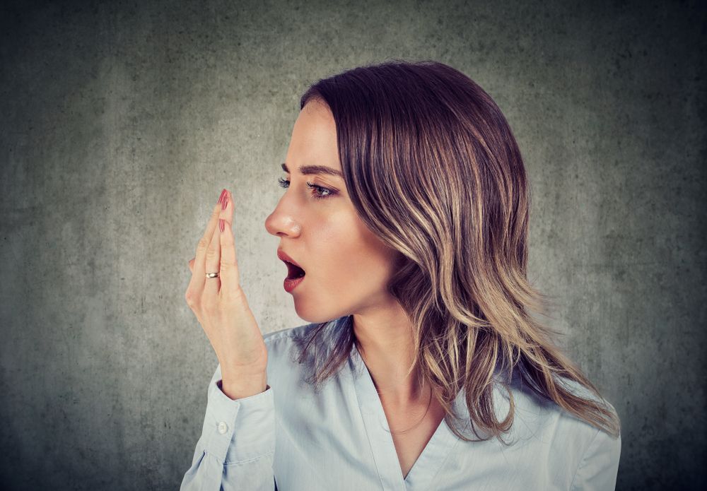 Woman checking her breath with hand in front of mouth, looking concerned.