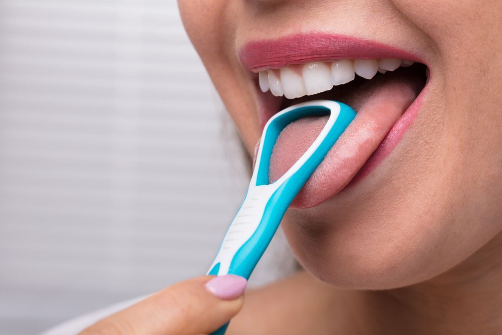 Woman using a blue and white tongue scraper to clean her tongue in a bathroom.