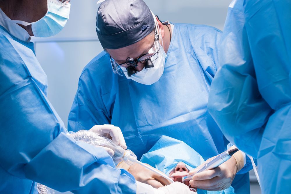 Surgeons in blue scrubs perform surgery on a patient in an operating room; overhead lighting.