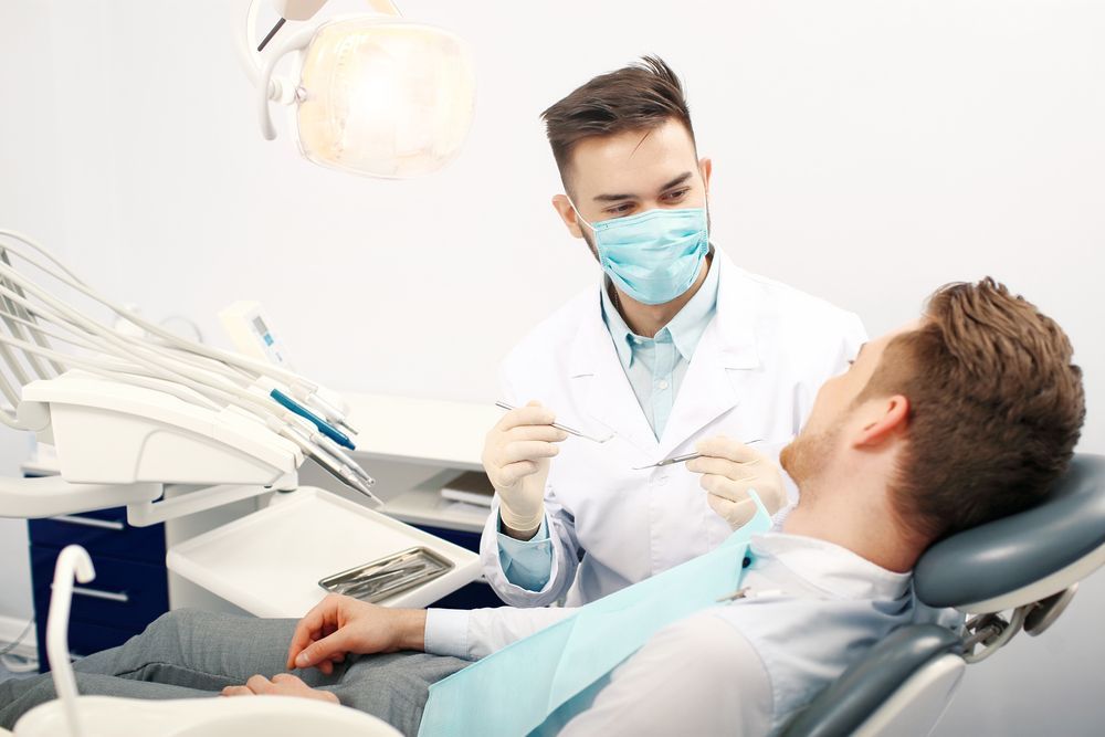 Dentist examining a patient's teeth in a dental chair; medical setting, wearing mask and gloves.