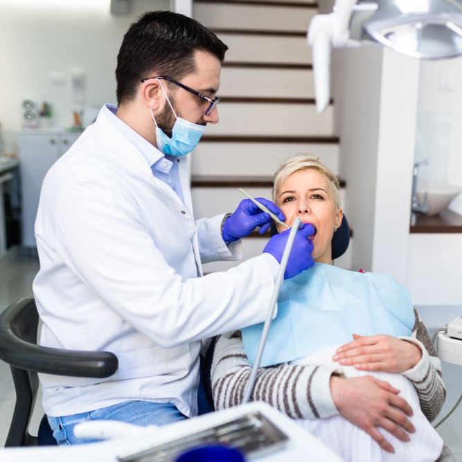 Dentist examining patient's teeth with tools in a dental office. The patient is sitting in a chair.