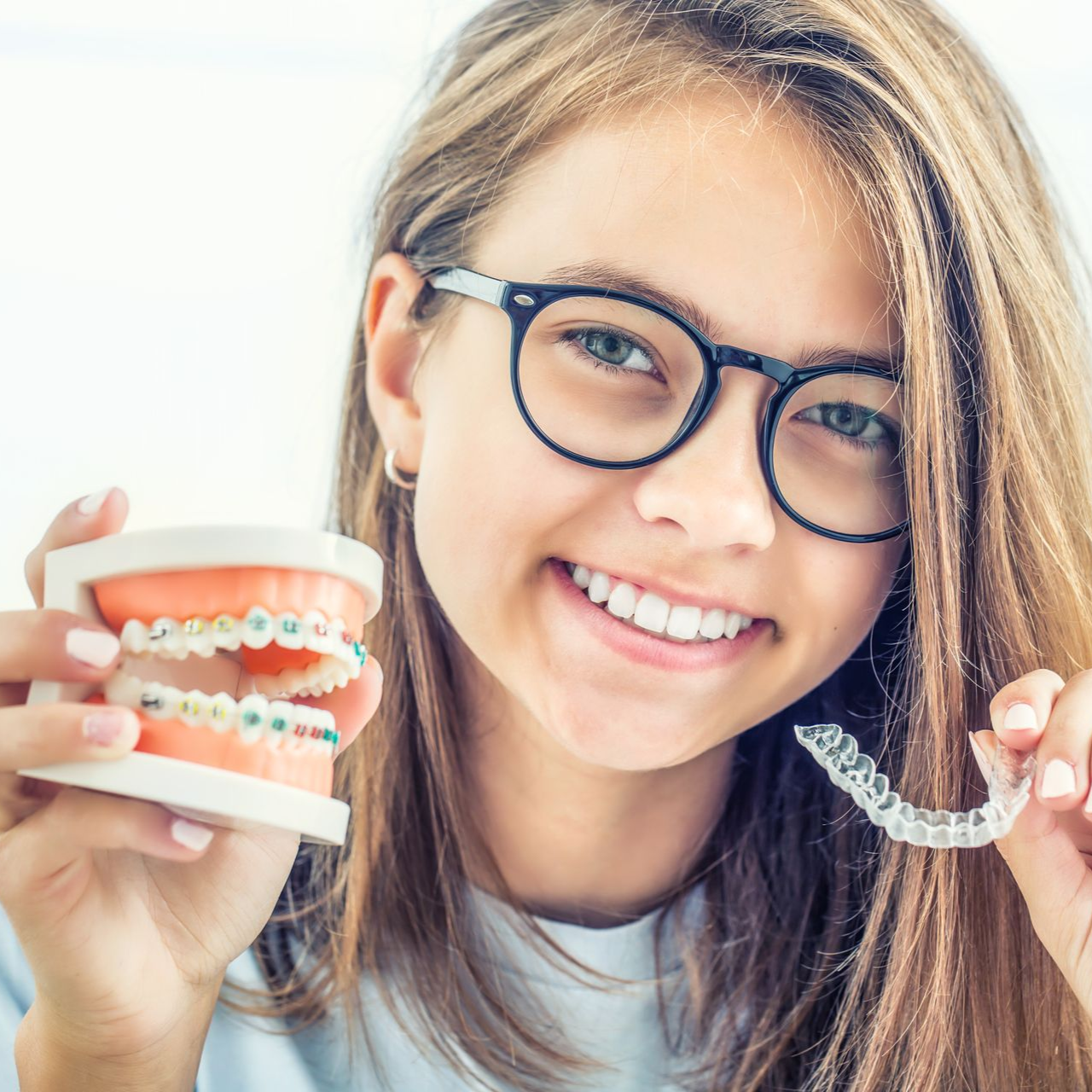 Girl with glasses smiling, holding orthodontic braces model and clear aligner.