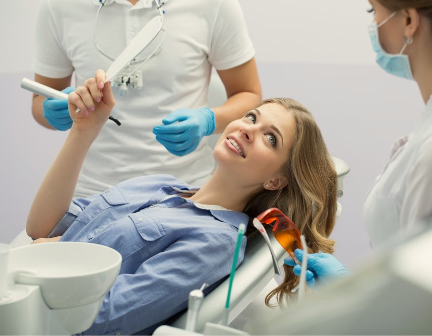 Woman smiles, looking at her teeth with a dental mirror, after a procedure. Two dental staff watch.