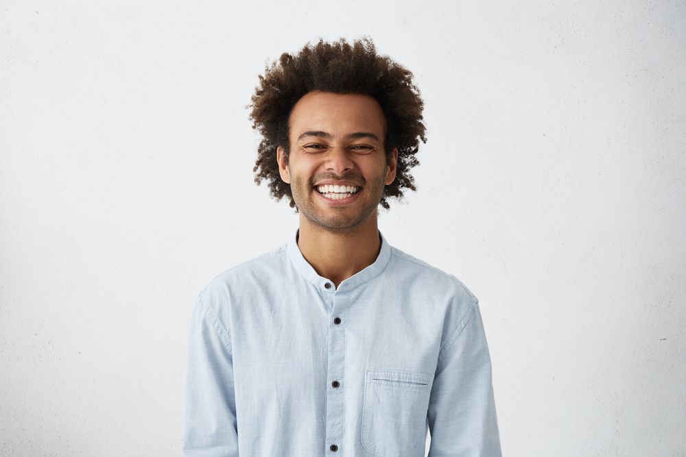 Man with afro smiling widely, wearing a light blue button-up shirt against a white backdrop.