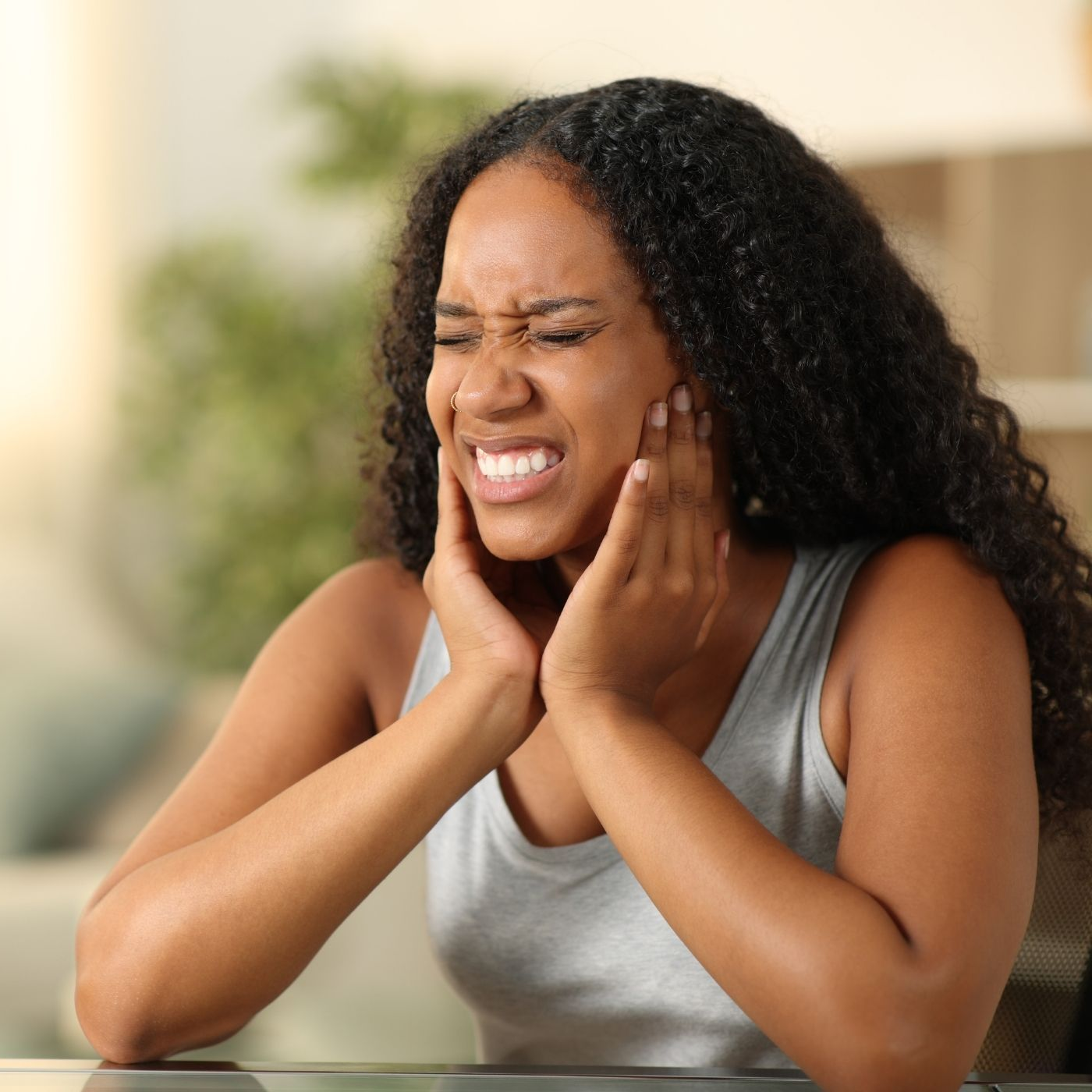 Woman with curly hair grimaces, holding her jaw in pain. Indoors.