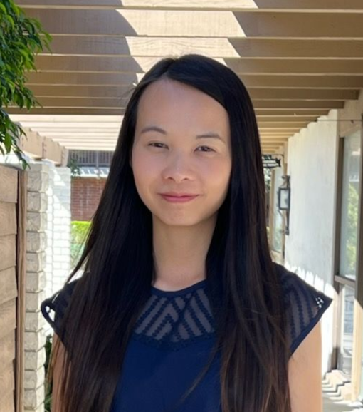 Woman with short brown hair smiles, wearing a white sweater, outdoors under a covered walkway.