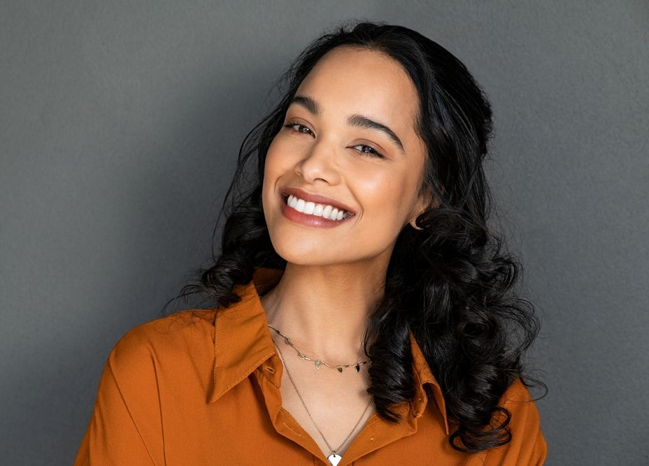 Woman with dark curly hair smiles, wearing an orange collared shirt and necklace against a gray backdrop.