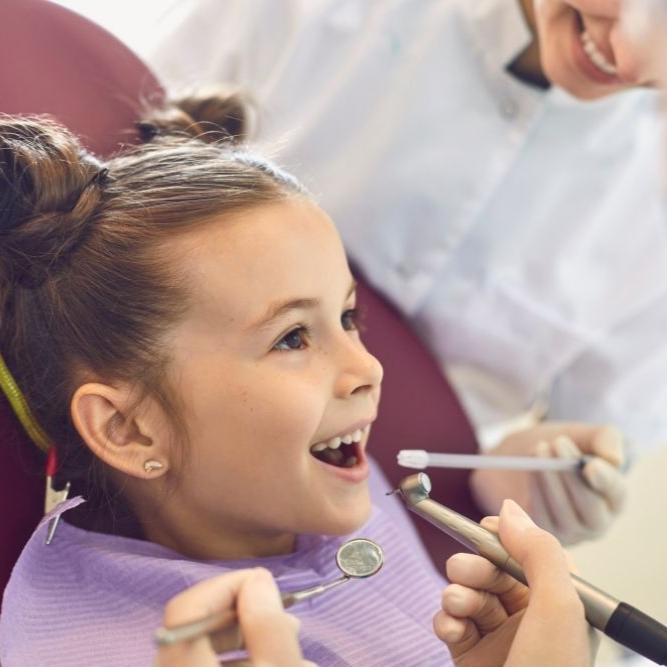 Child in dentist chair, smiling with mouth open; dental tools and dentist visible.