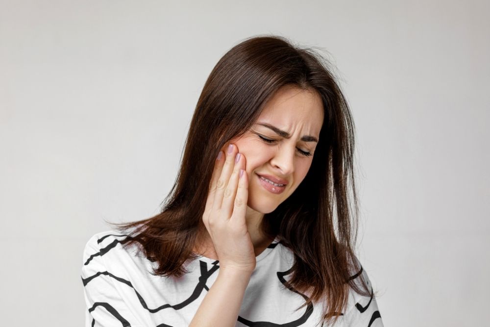 Woman with long brown hair, clutching her cheek in pain, eyes closed, suffering toothache.