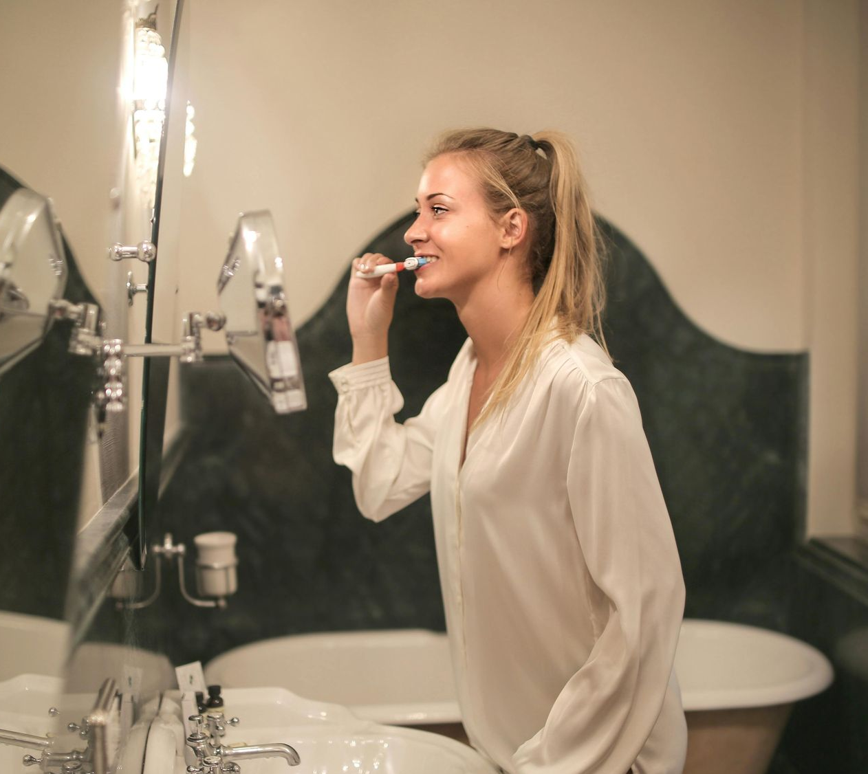 Woman brushing teeth in bathroom, looking at mirror; white shirt, ponytail.