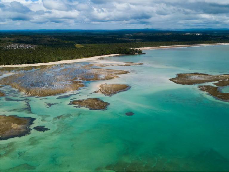 Águas costeiras azuis com afloramentos rochosos, uma praia de areia e um litoral arborizado sob um céu nublado. Pousada Vila da Barra