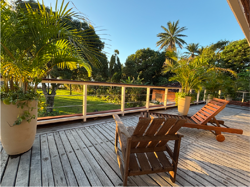 Deck de madeira com espreguiçadeiras e palmeiras em vasos com vista para um quintal ensolarado com árvores. Pousada Vila da Barra