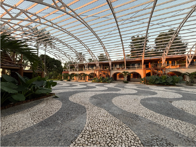 Praça de paralelepípedos com padrão ondulado e telhado de treliça arqueada, levando a um edifício com arcos e plantas. Pousada Vila da Barra