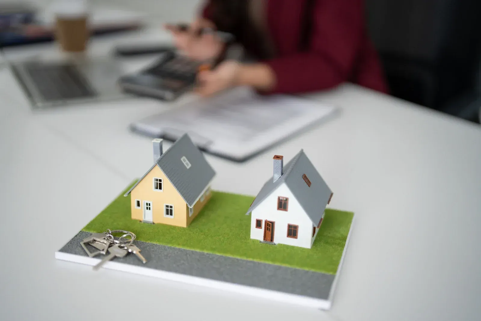Miniature houses and keys on a table, a person blurred in the background working with documents.